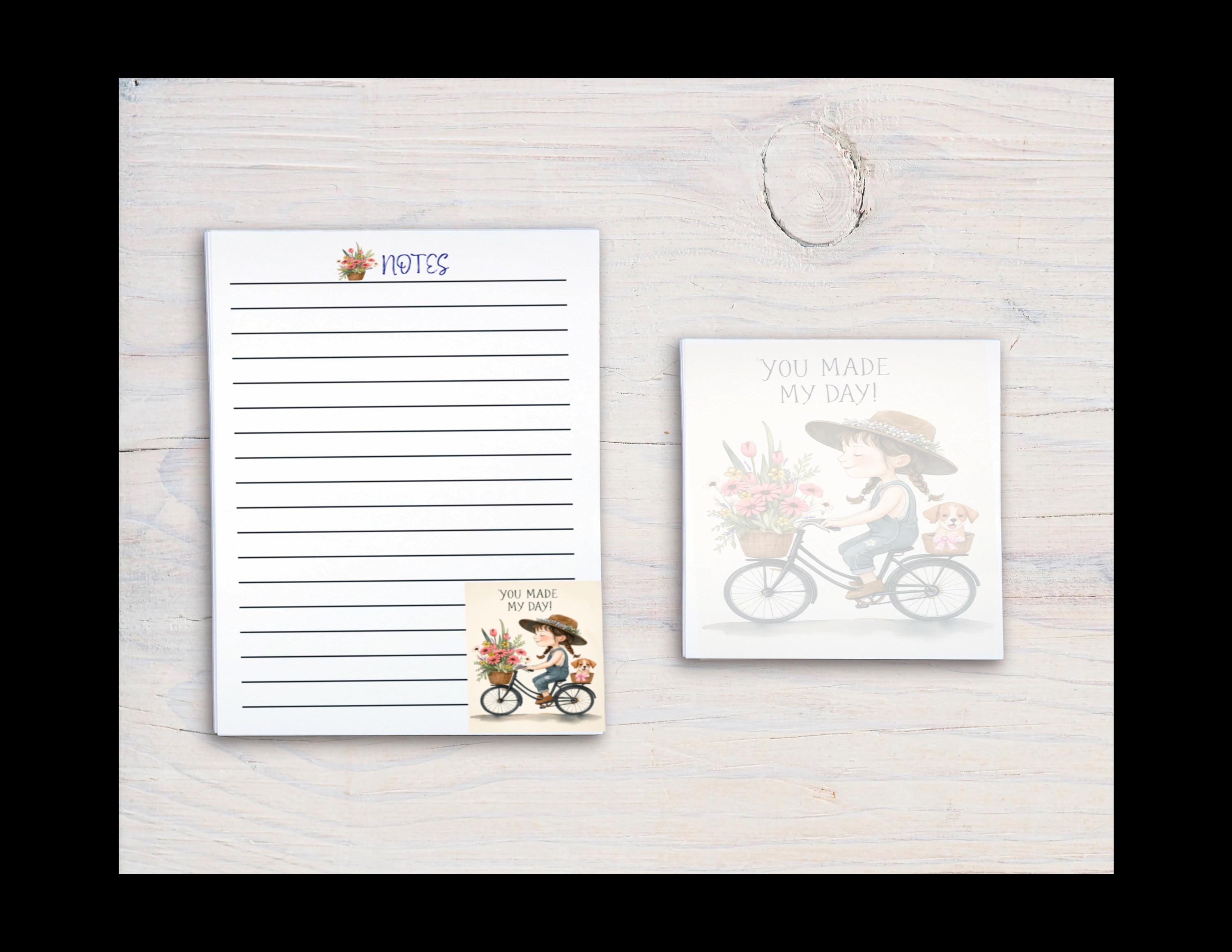 a white wooden surface with two items placed on it: a lined paper pad and a small card with a bicycle and flowers.