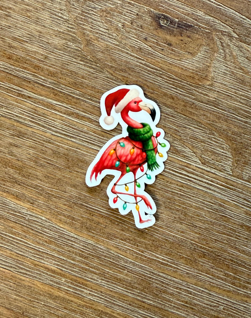A sticker of a flamingo wearing a Santa hat and scarf, with Christmas lights around its feet, is placed on a wooden surface.