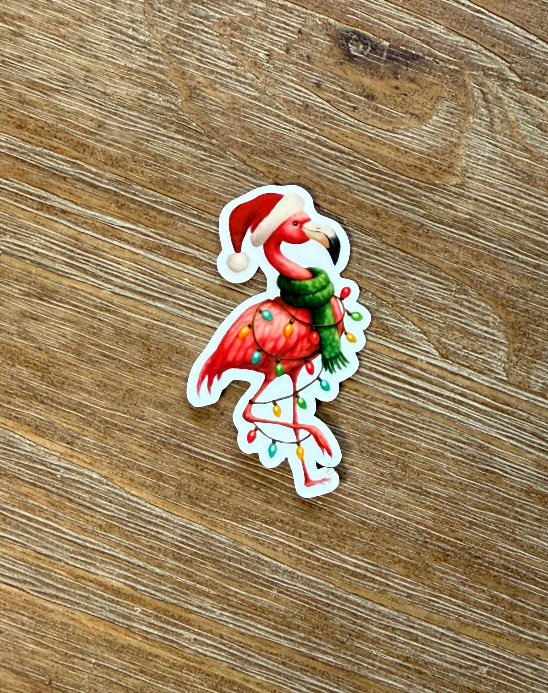 A sticker of a flamingo wearing a Santa hat and scarf, with Christmas lights around its feet, is placed on a wooden surface.