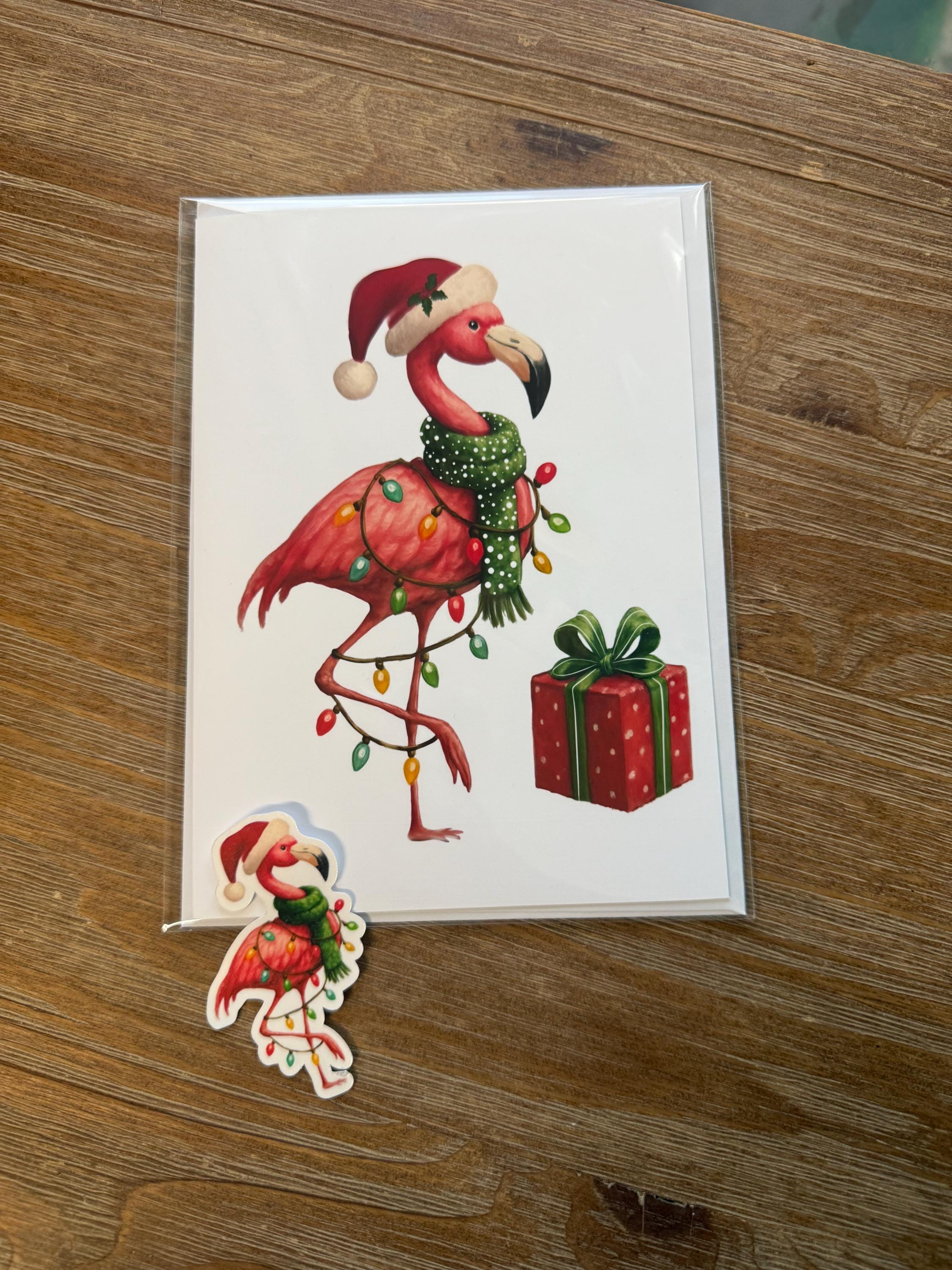 A greeting card featuring a pink flamingo wearing a Santa hat and holding a gift box, along with a small Christmas-themed sticker.