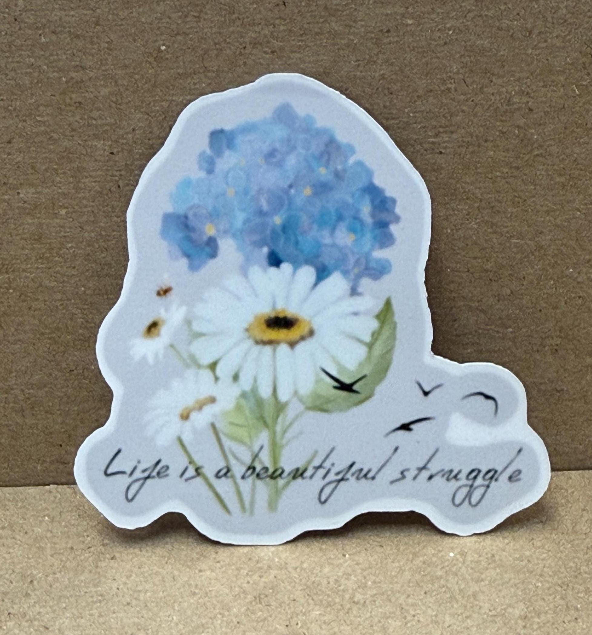 A sticker with a blue flower and white daisies, along with the text &quot;Life is a beautiful struggle&quot; written below it.