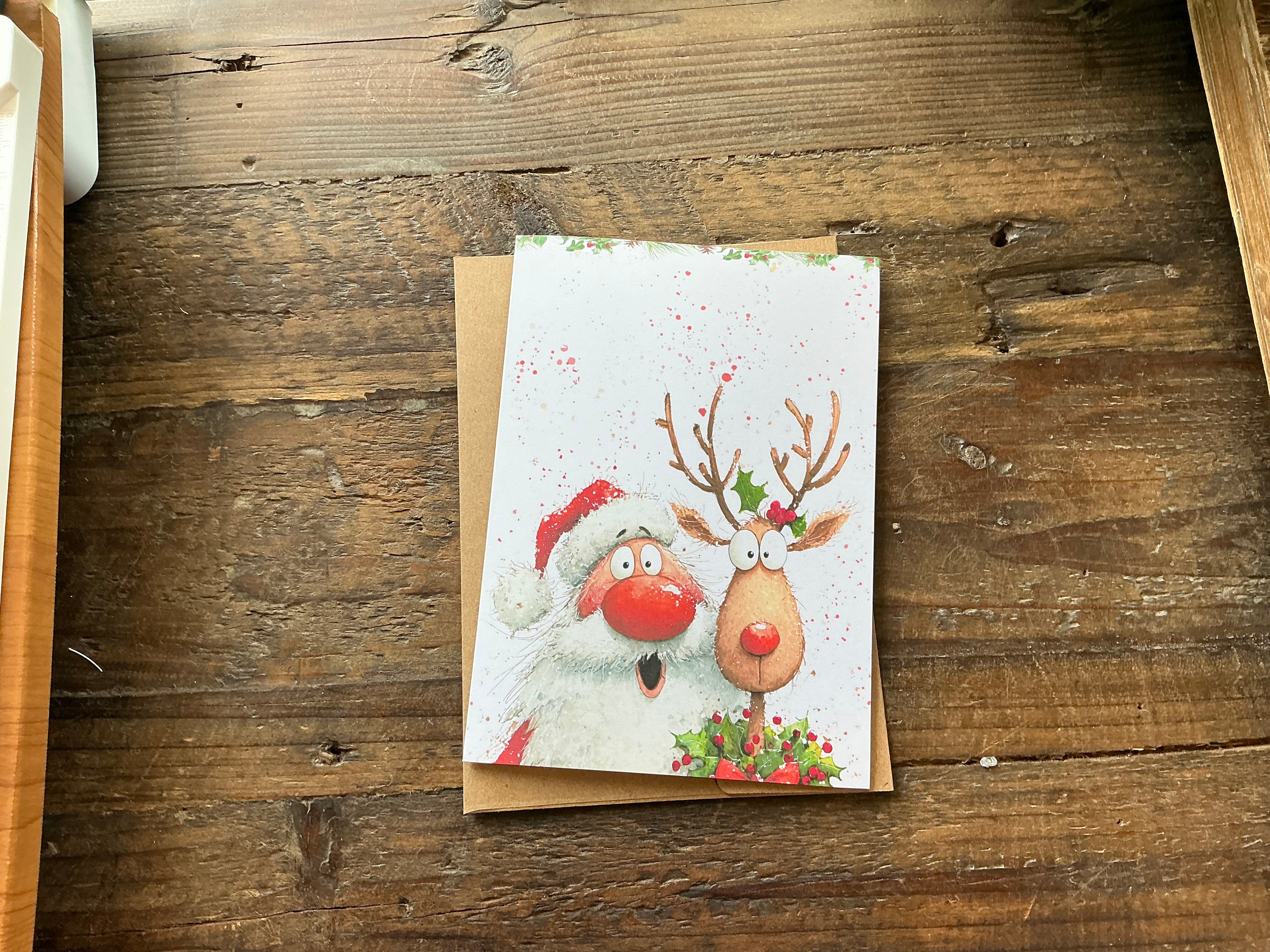 A Christmas card featuring a Santa Claus and a reindeer, with the Santa wearing a red hat and the reindeer wearing a red nose, sitting on a wooden surface.