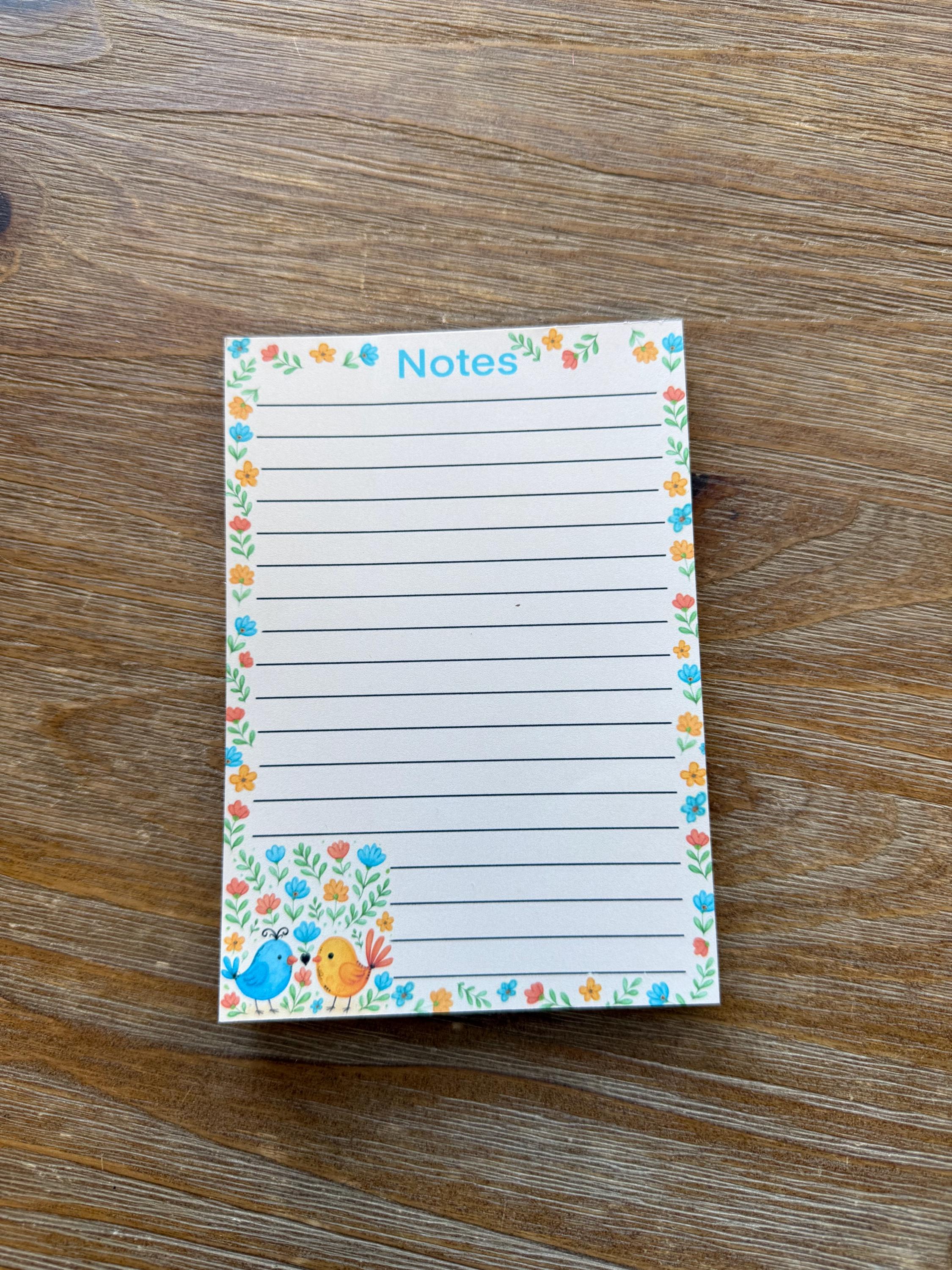 A lined notepad with a floral border and a small illustration of a bird on the right side, placed on a wooden surface.