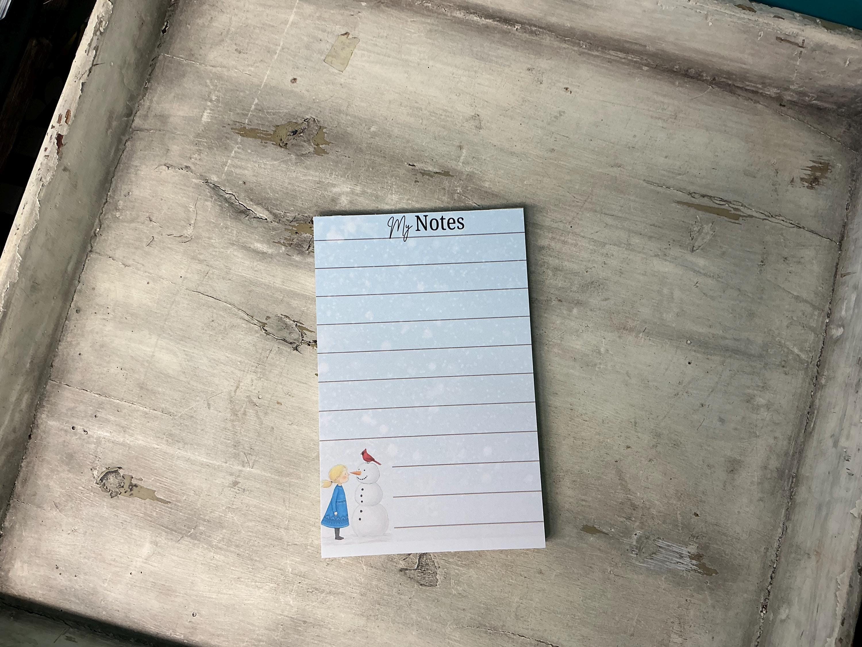A lined notepad with a snowman illustration on the cover, placed on a wooden surface.