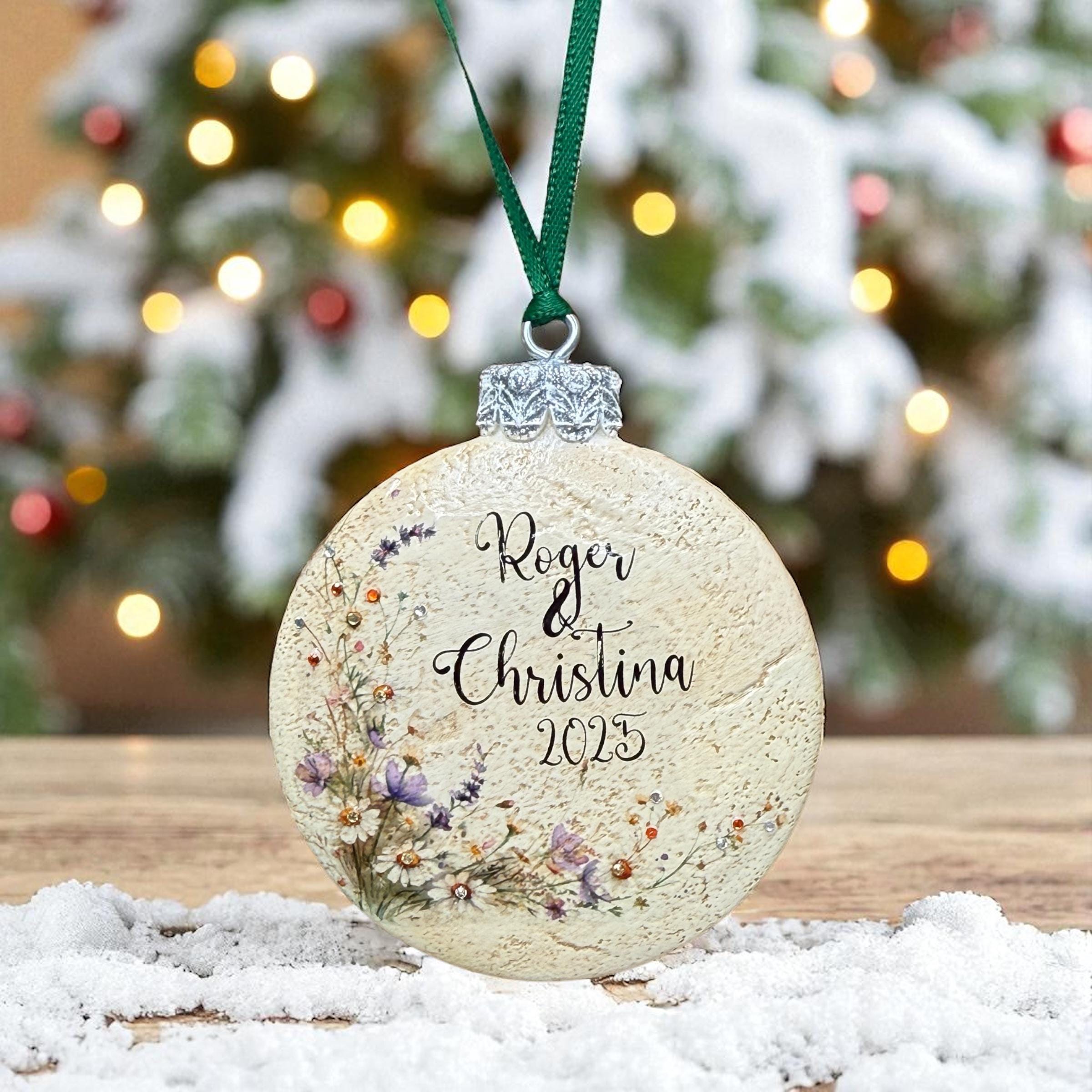 A round ornament with a floral design and the names "Rogers & Christina" written on it, hanging from a green ribbon.