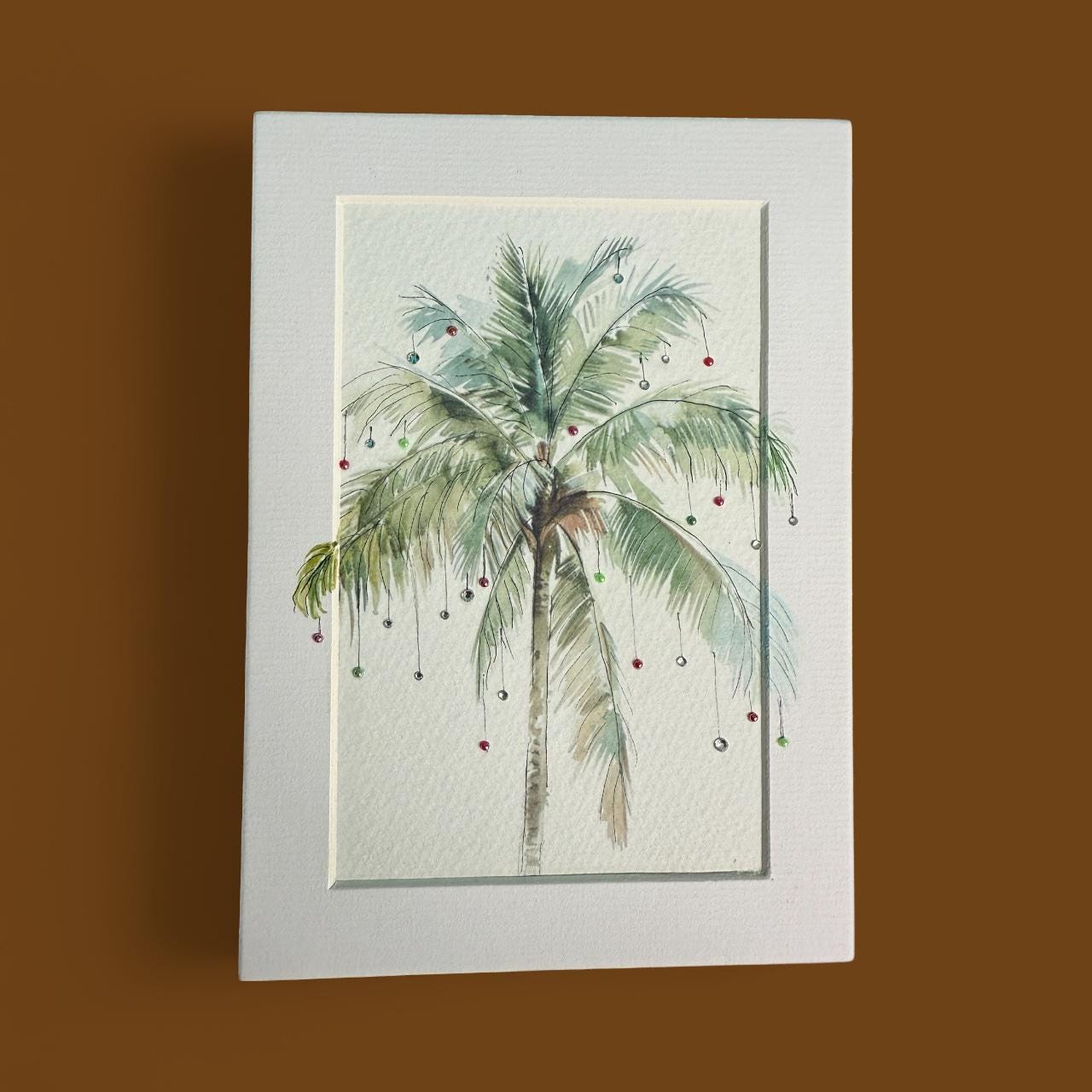 A framed watercolor painting depicting a palm tree with green leaves and red ornaments hanging from its branches.
