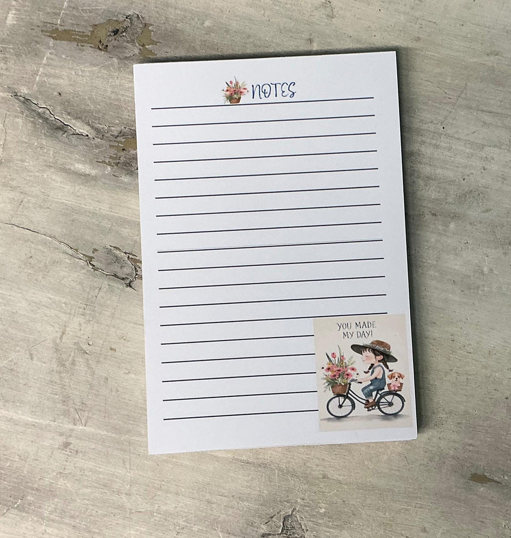A blank lined notepad with a sticker featuring a person riding a bicycle and flowers, placed on a wooden surface.