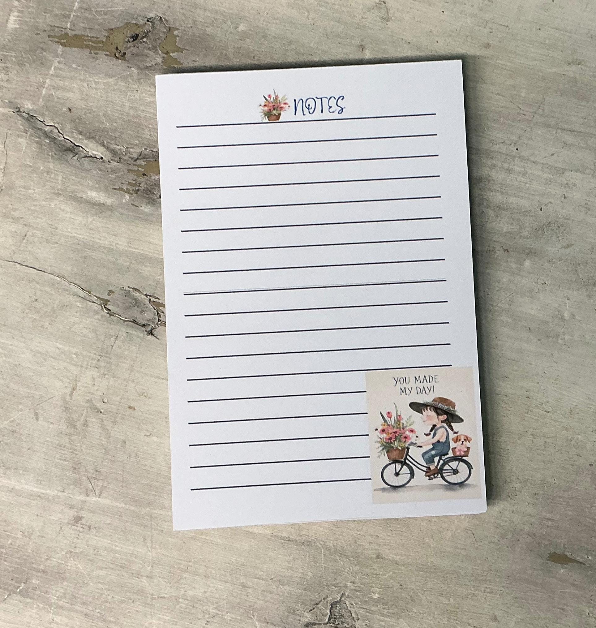 A blank lined notepad with a sticker featuring a person riding a bicycle and flowers, placed on a wooden surface.