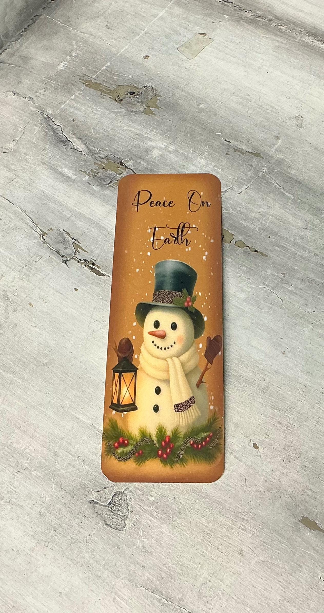 A snowman holding a lantern and surrounded by snow and greenery, with the text &quot;Peace on Earth&quot; written on it.