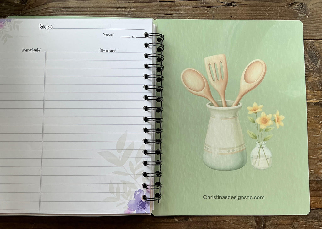 an open spiral-bound recipe book with a page displaying an illustration of a vase with flowers and wooden spoons.