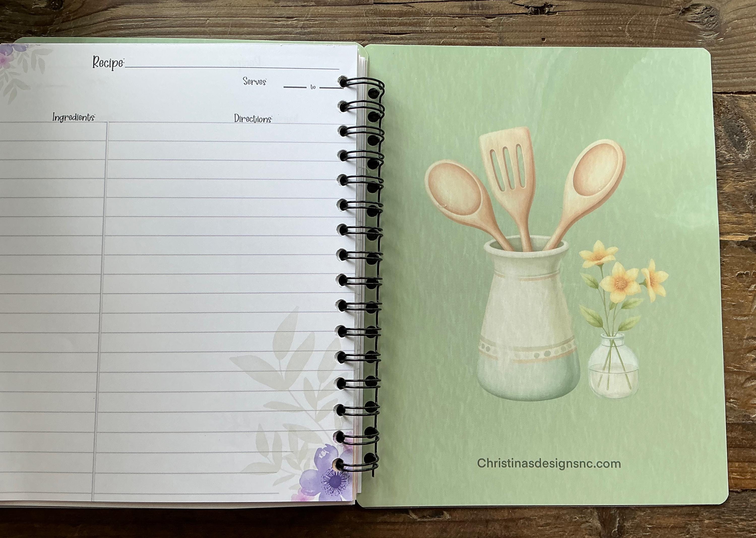 an open spiral-bound recipe book with a page displaying an illustration of a vase with flowers and wooden spoons.