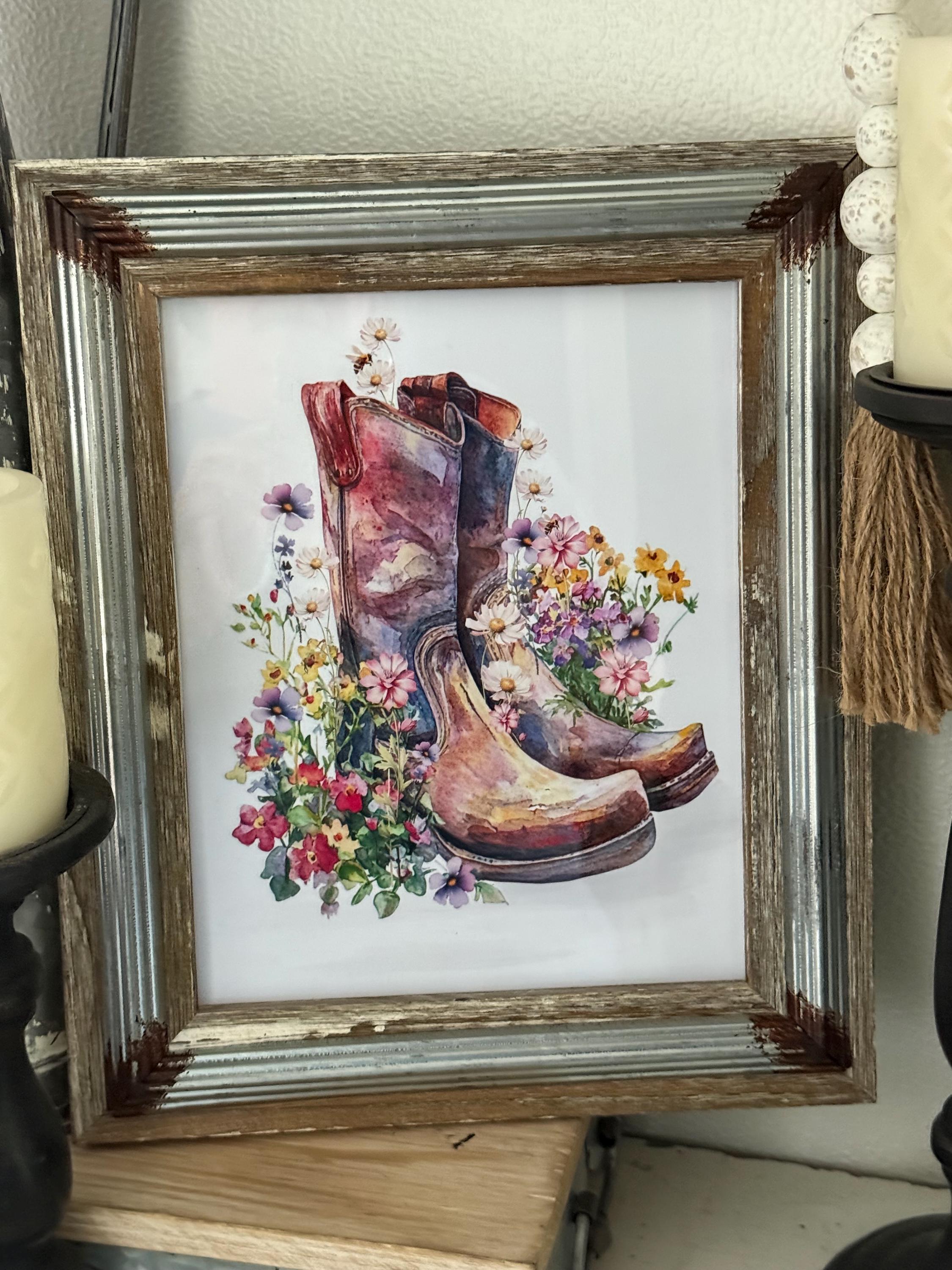 A framed print depicting a pair of old, worn cowboy boots surrounded by a vibrant floral arrangement.