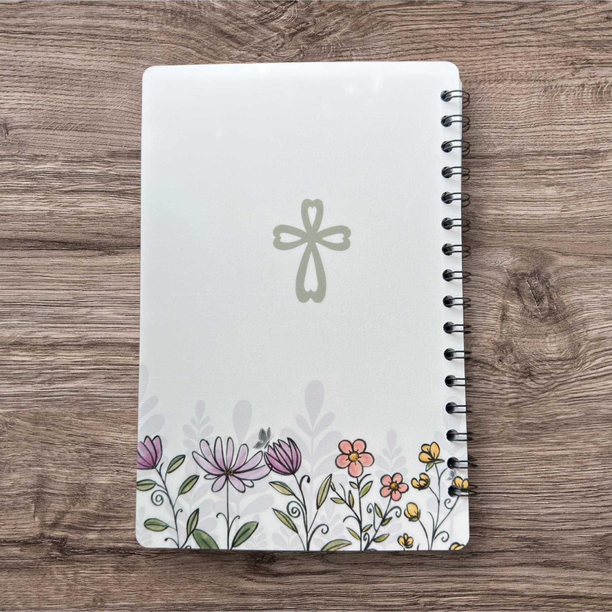 A white spiral notebook with a floral design on the cover, featuring a cross and various colorful flowers.