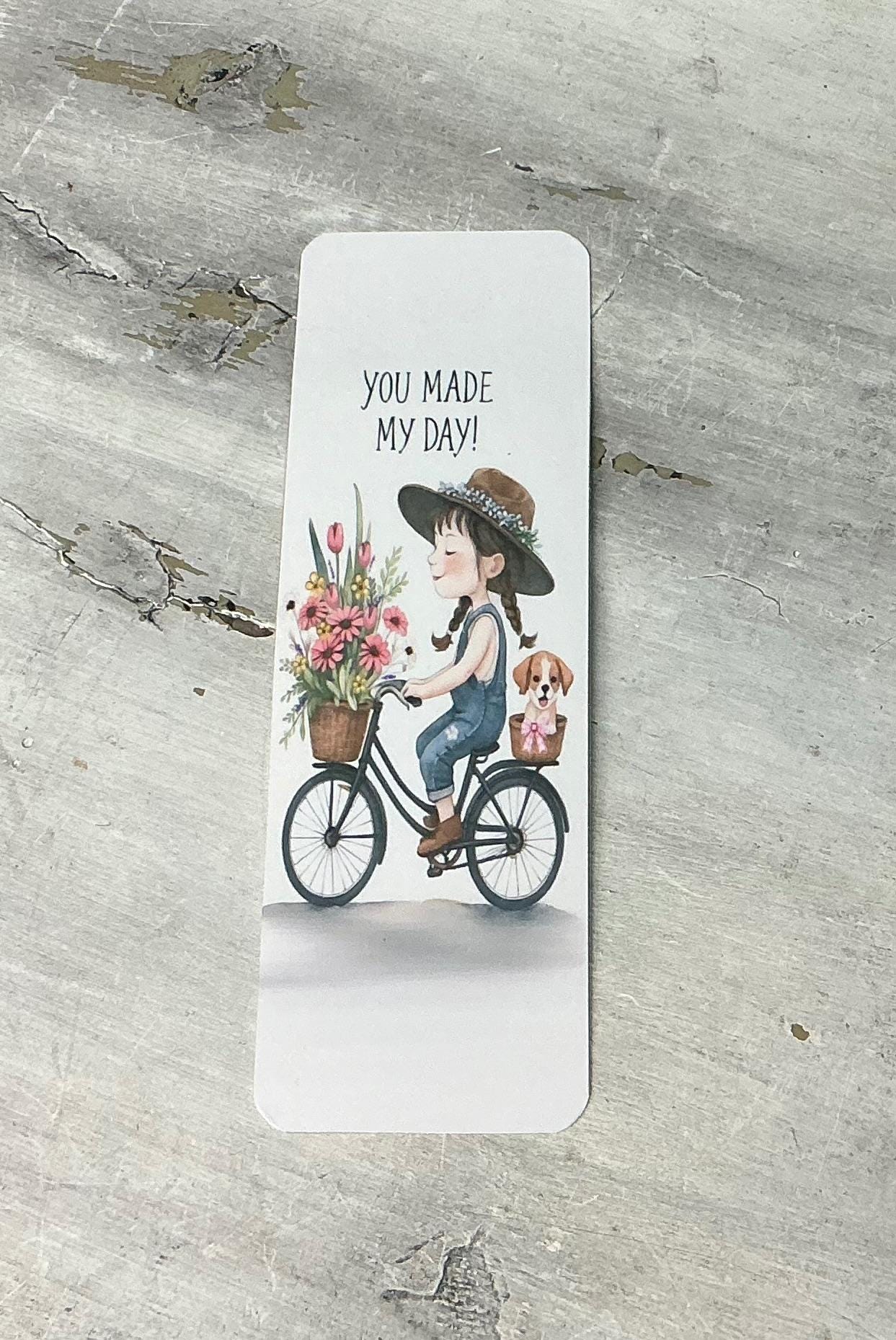 A white bookmark with an illustration of a young girl riding a bicycle with a basket of flowers, accompanied by the text &quot;You made my day!&quot;