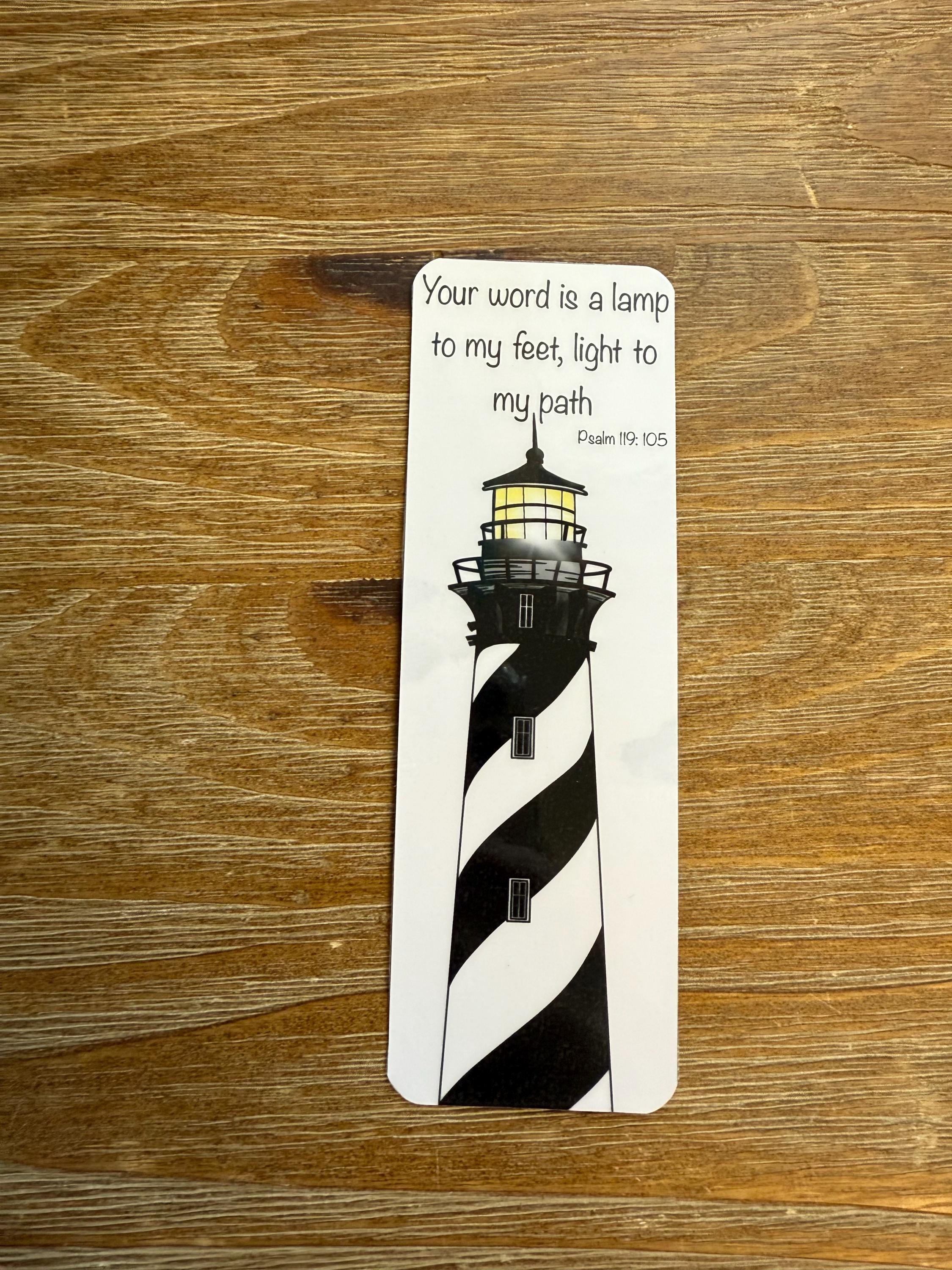 A white rectangular bookmark with a black and white lighthouse illustration on it, placed on a wooden surface.
