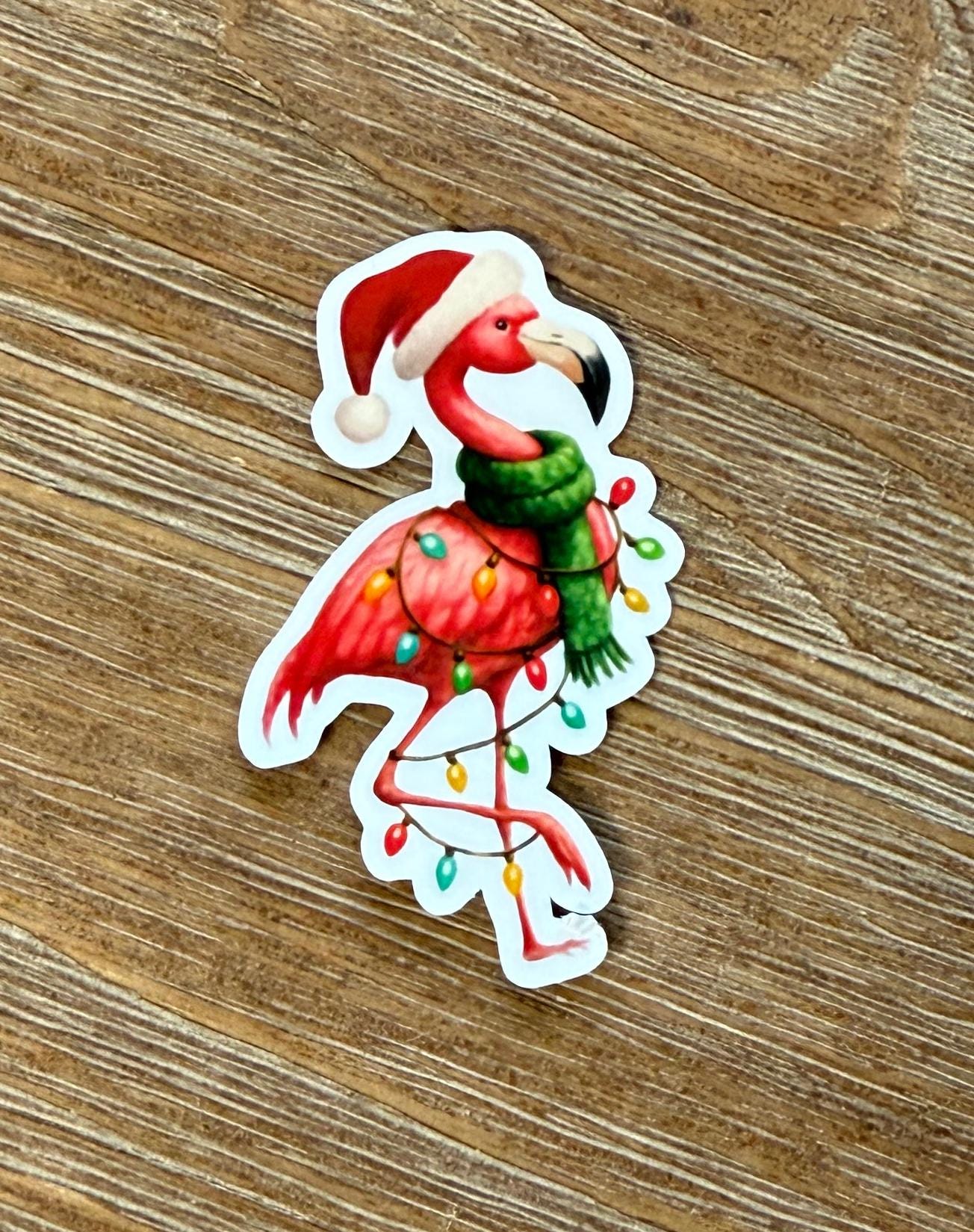 A sticker depicting a flamingo wearing a Santa hat and scarf, with Christmas lights around its neck.