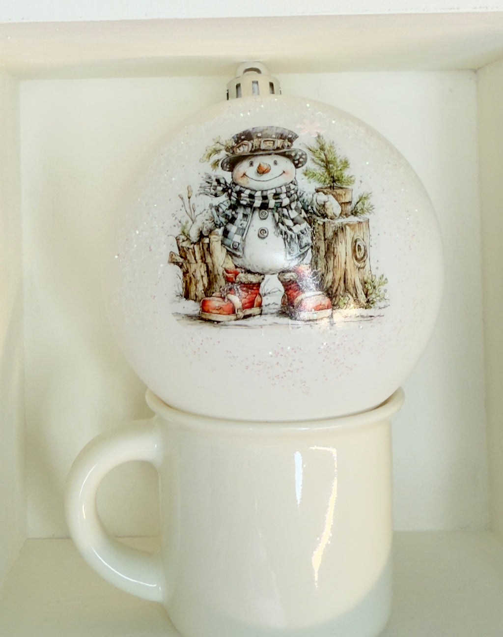 A white mug with a snowman ornament on it, placed on a shelf.