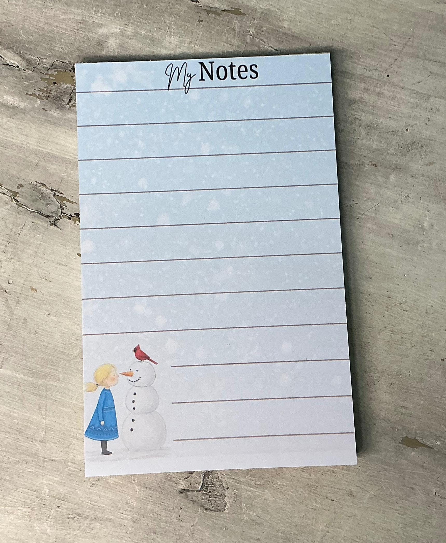 A lined notepad with a snowman and a girl on it, placed on a wooden surface.