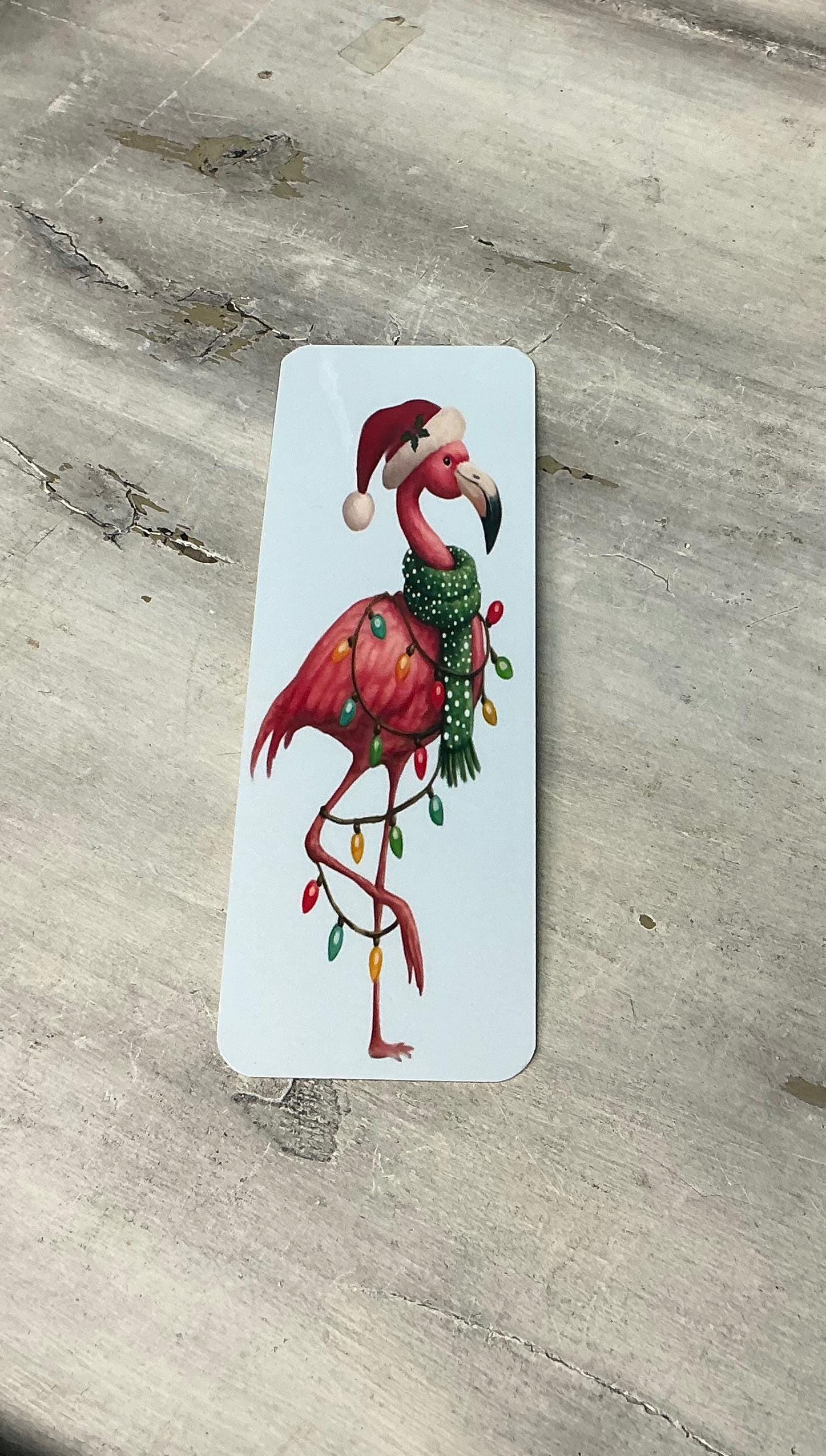 A pink flamingo wearing a Santa hat and scarf is standing on one leg on a wooden surface.