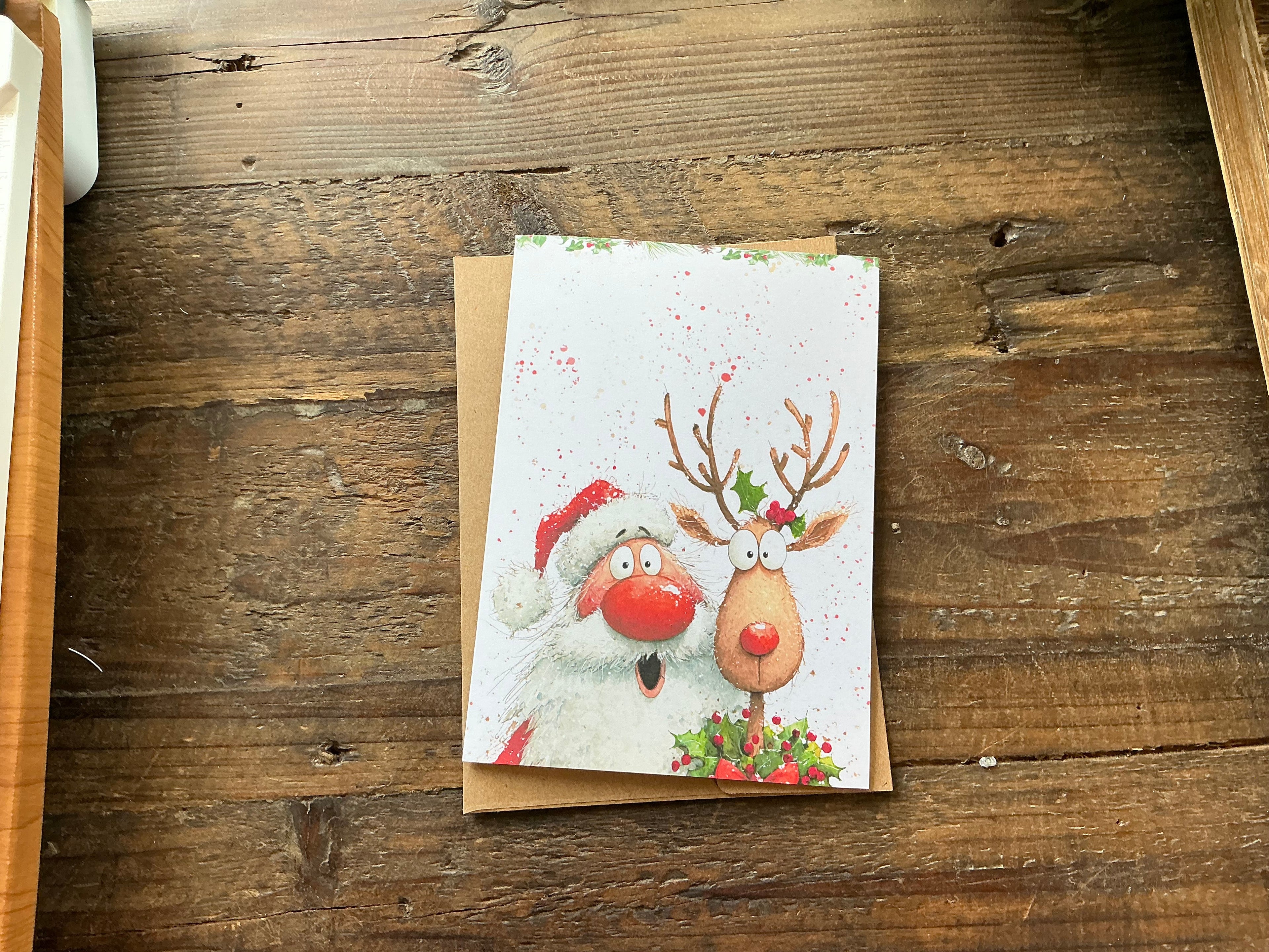 A Christmas card featuring a Santa Claus and a reindeer, with the Santa wearing a red hat and the reindeer wearing a red nose, sitting on a wooden surface.