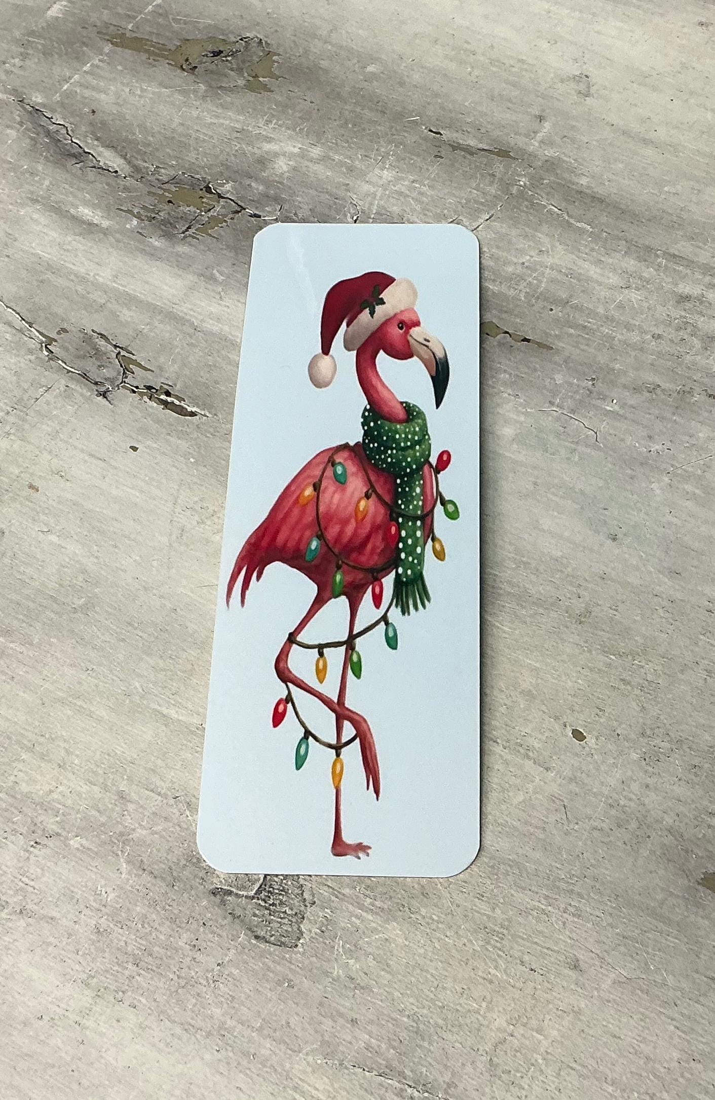 A pink flamingo wearing a Santa hat and scarf with Christmas lights is standing on a gray surface.