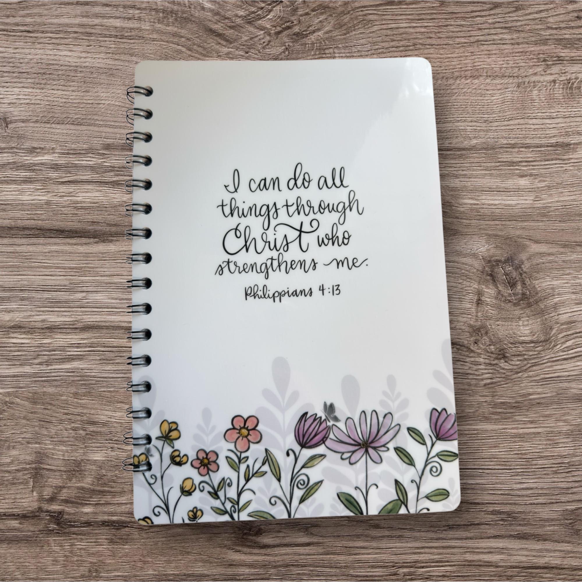 A spiral-bound notebook with a floral design and a Bible verse printed on the cover.