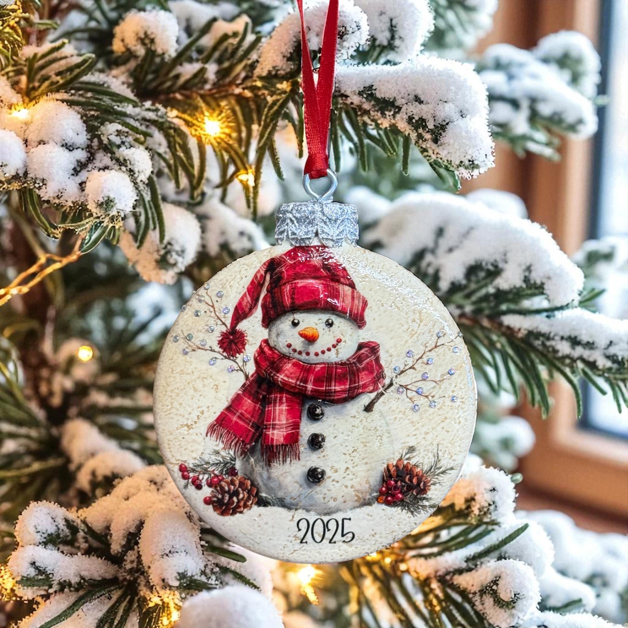 A snowman ornament with a red scarf and hat is hanging from a tree branch, surrounded by snow-covered branches and twinkling lights.